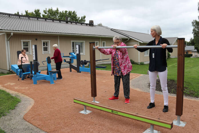 Vapaa-aikapäällikkö Emilia Andersson näyttää Eila Paloselle, miten tangosta saa tukea vaikkapa kyykkyharjoituksiin. Taustalla ulkokuntosaliin ovat tutustumassa myös Helka Koivula ja Kaija Saario.